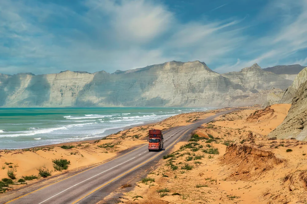 Truck traveling along the Makran Coastal Highway in Balochistan, with rugged cliffs and the Arabian Sea coastline in the background [Image via Getty Images].