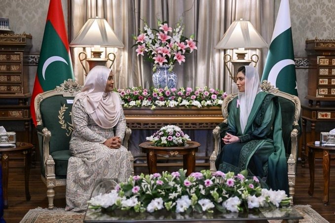 The two First Ladies seated side by side in a reception room between the flags of both countries.